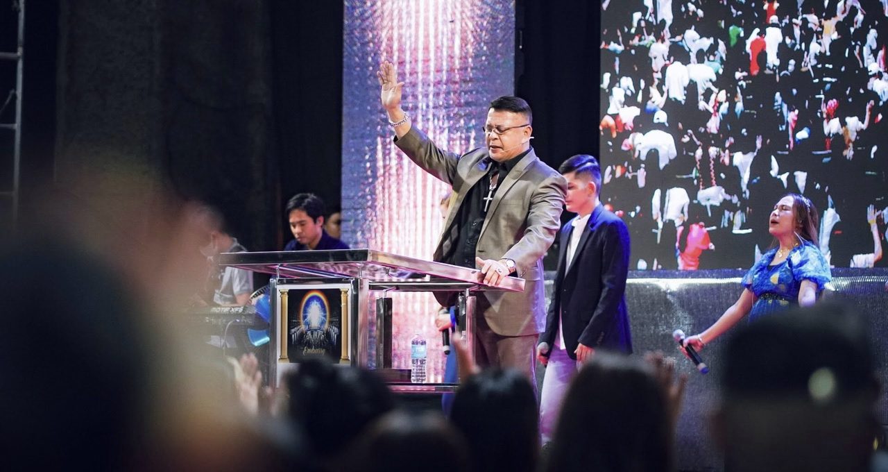 Bishop Rod Cubos with his hand raised in prayer at the pulpit, the congregation shown on the screen behind him